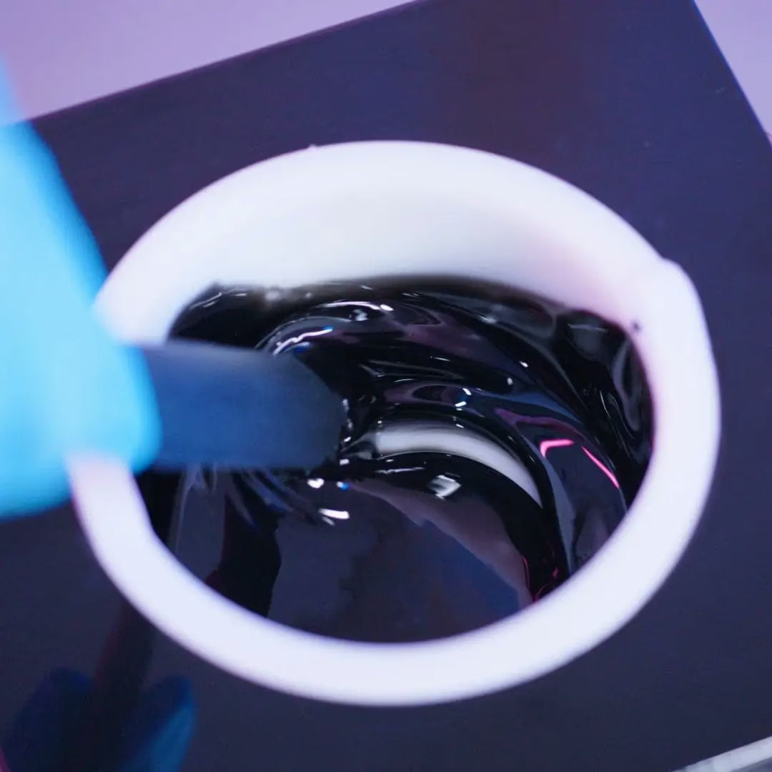 A person wearing a blue glove stirs a melted Black Coral Hard Wax by Black Coral Wax with a black spatula. The thick, shiny liquid swirls and reflects light, against a dark, blurred background.