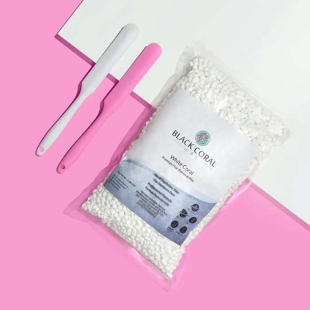 A 5lb Warmer + FREE Hot Wax from Black Coral Wax, filled with white wax beads, is shown on a pink and white background beside two waxing spatulas—one white and one pink.