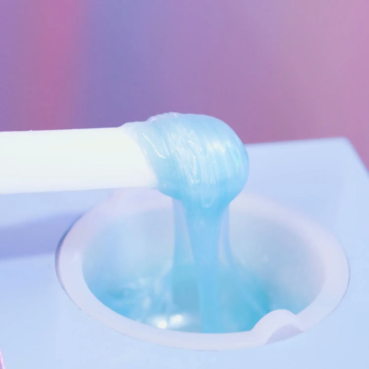A close-up of Black Coral Wax  Kai Hard Wax, translucent blue and ideal for sensitive skin, being dispensed from a white tube into a round container against a softly blurred pink and purple backdrop.
