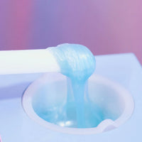 A close-up of Black Coral Wax  Kai Hard Wax, translucent blue and ideal for sensitive skin, being dispensed from a white tube into a round container against a softly blurred pink and purple backdrop.
