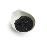 The Boss Babe Wax Glittera by Black Coral Wax USA is showcased in a white ceramic bowl akin to a Black Coral Hard Wax vessel. The bowls subtle, spout-like design elegantly cradles small, shiny black beads against a plain backdrop, reflecting potential for long-lasting hair removal.
