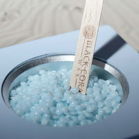 Blue kai  hard wax wax beads in a container with a wooden spatula labeled 'Black Core Wax'.
